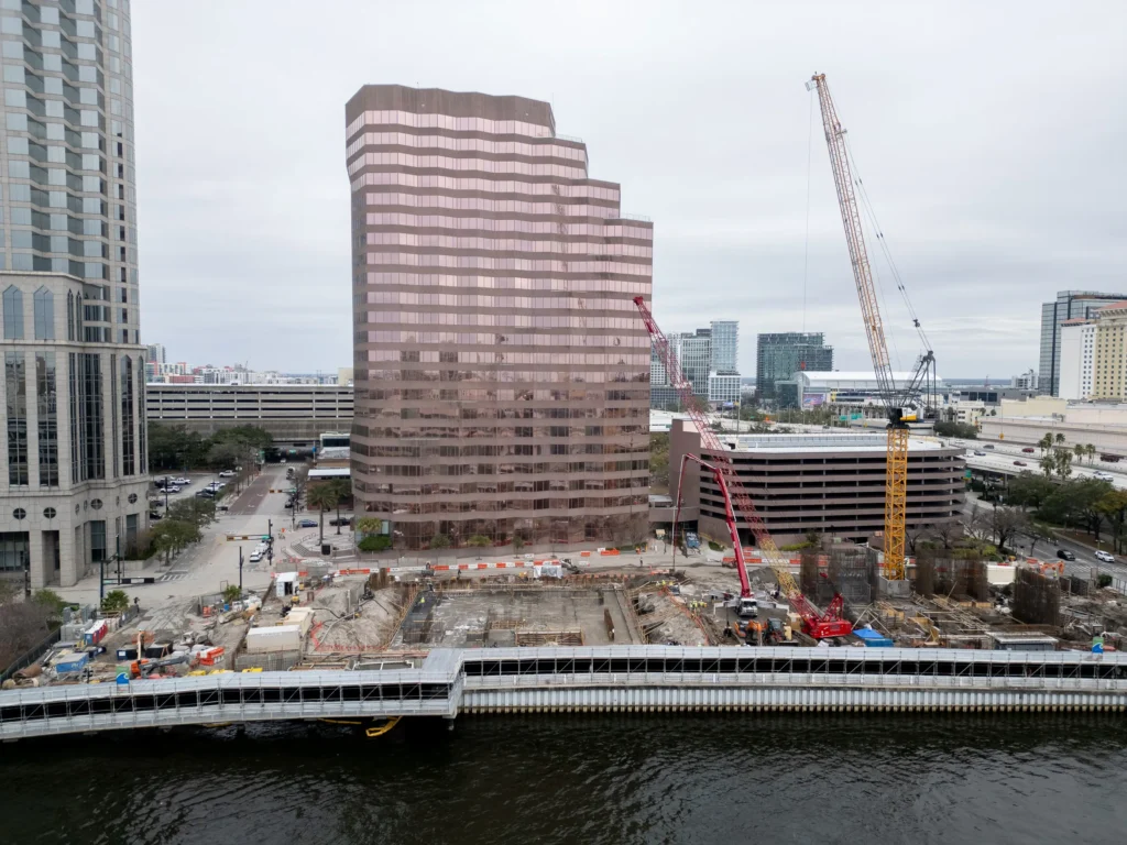 Aerial view of the Pendry Tampa construction site on the riverfront beside a rose-colored office tower, by Drone Permission