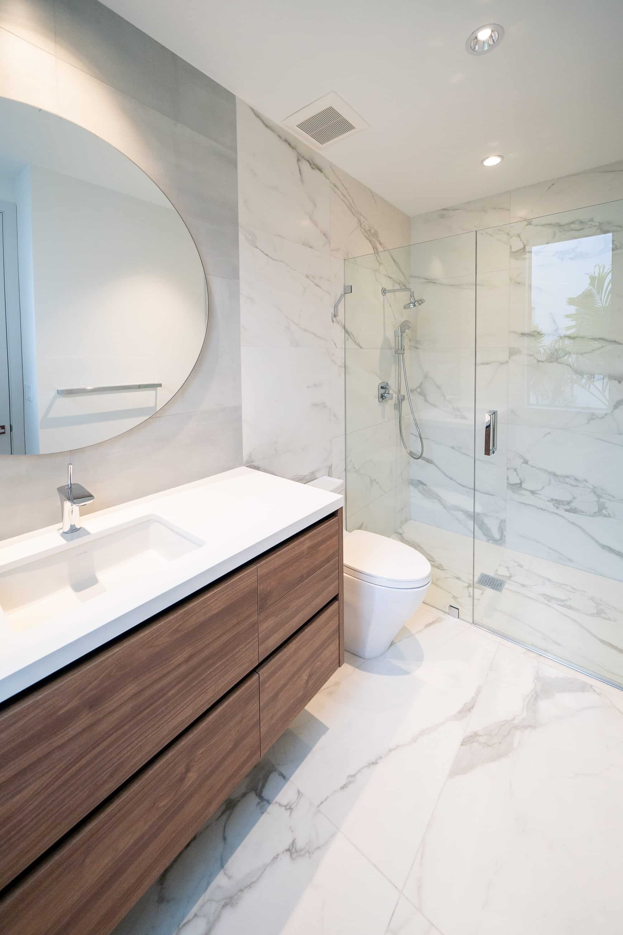 Modern bathroom with marble tile walls, round mirror, floating wood vanity and glass walk-in shower in a Delray Beach condo