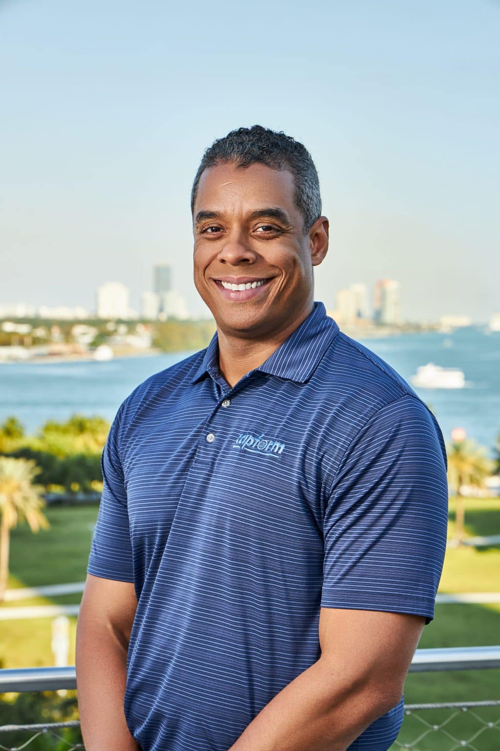 Outdoor corporate headshot of a smiling man in a navy polo with the Miami waterfront behind him