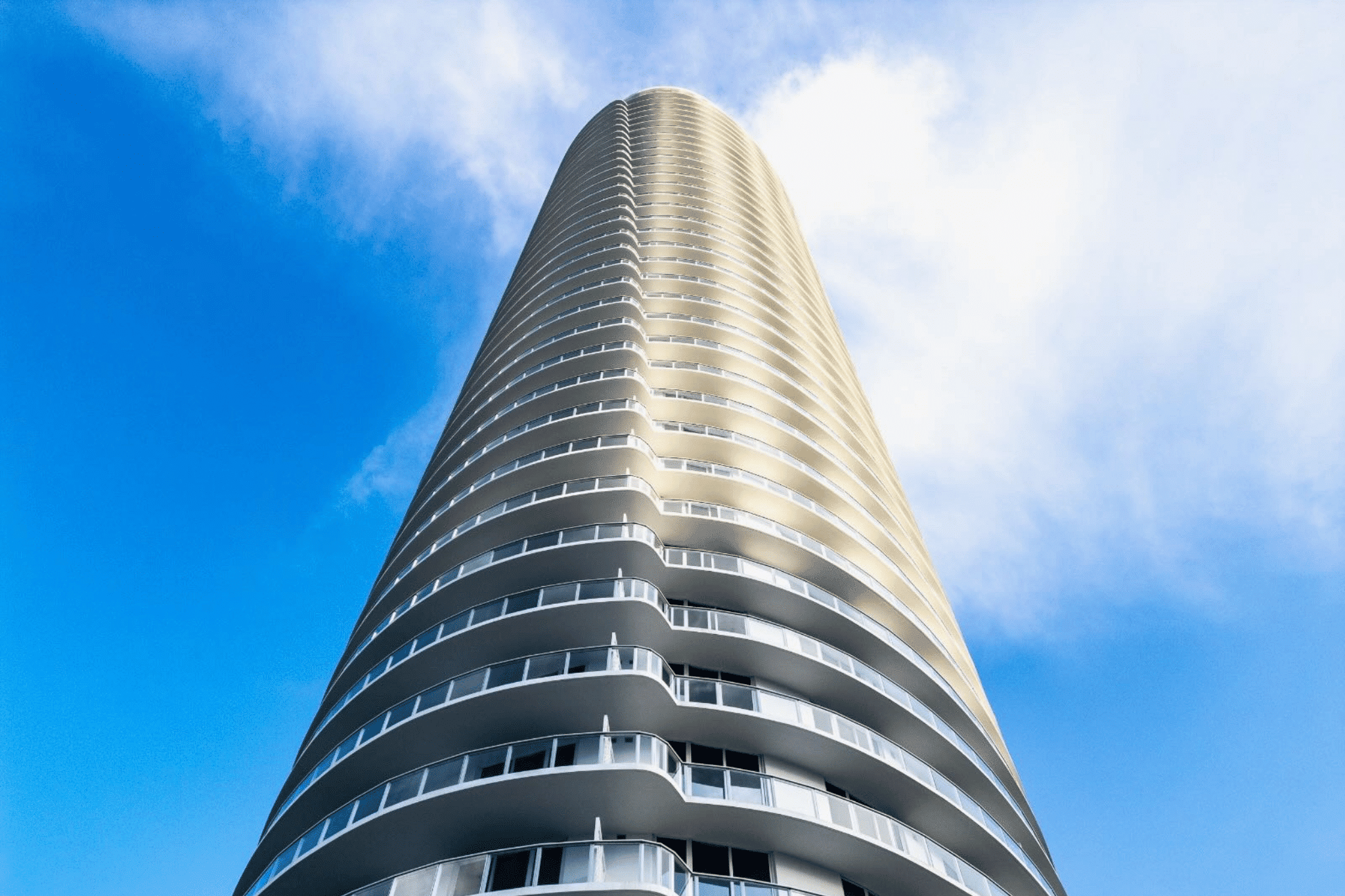 Low-angle view of a curved residential high-rise with rows of balconies against a blue sky in Miami