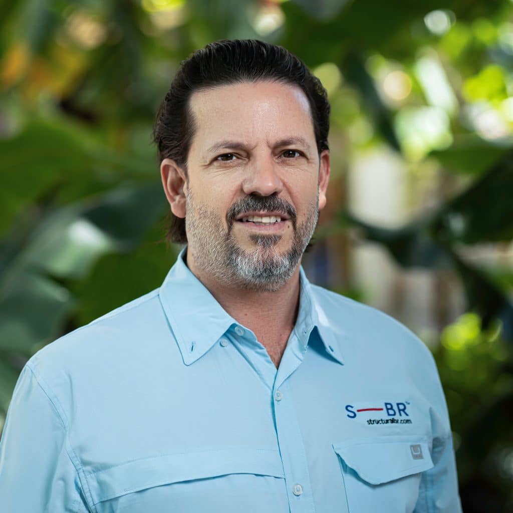 Outdoor headshot of a smiling bearded man in a light blue company shirt against tropical greenery