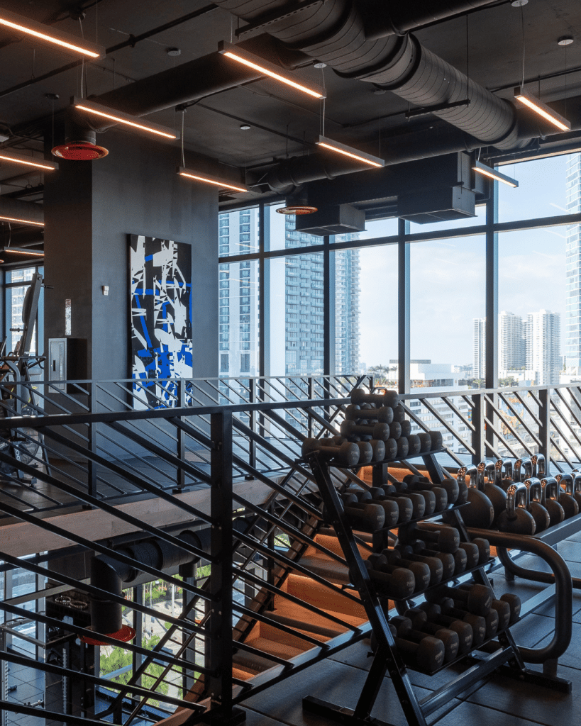 Modern high-rise gym with dumbbell racks and floor-to-ceiling windows overlooking a Florida city skyline