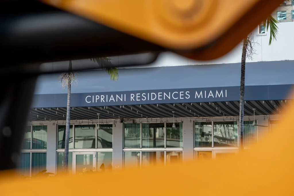 Cipriani Residences Miami lettering on a striped awning framed between palm trees, photographed past yellow construction equipment in the foreground