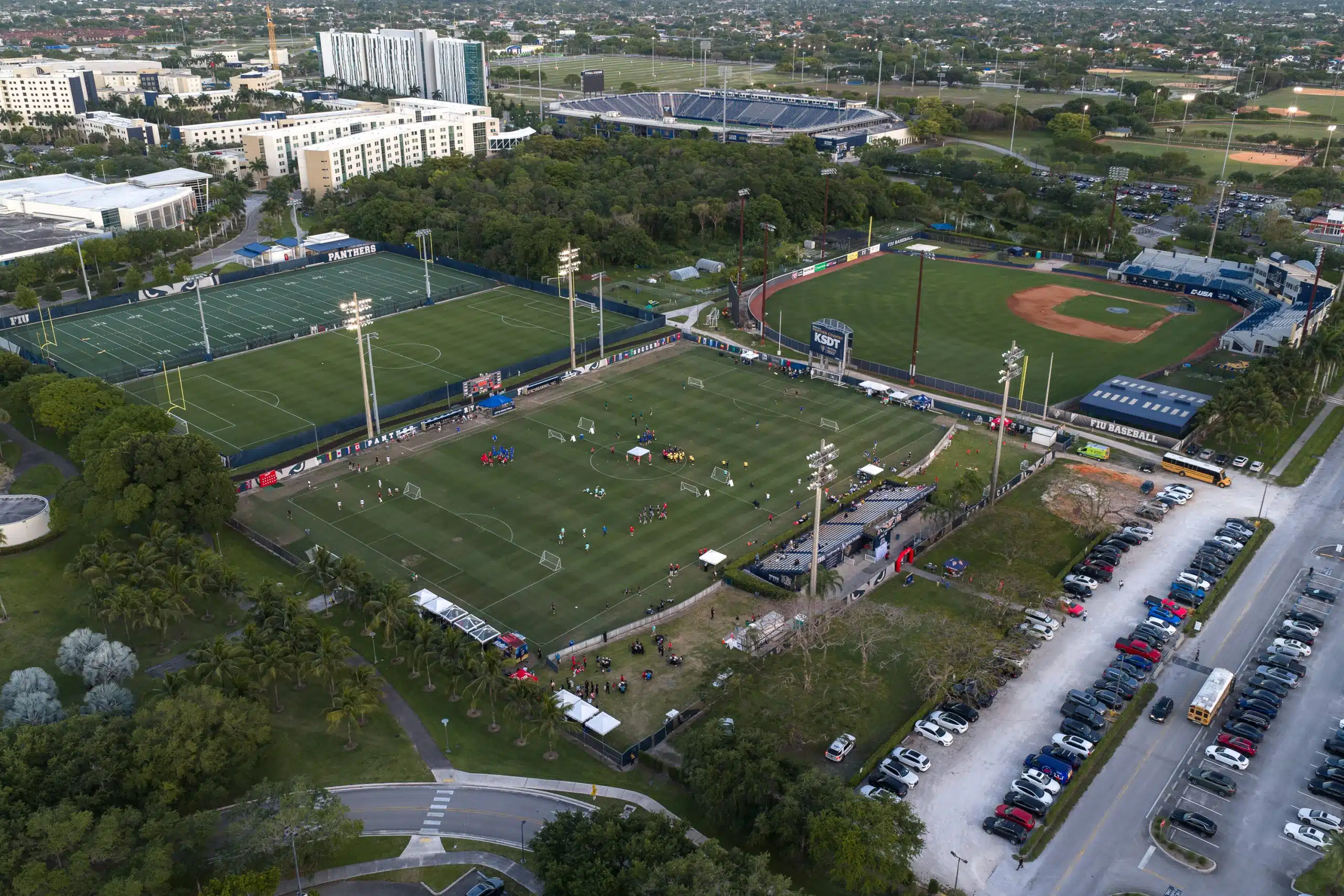 Evening aerial view of a soccer tournament in progress on the FIU campus fields in Miami, with the football stadium and baseball park in the background