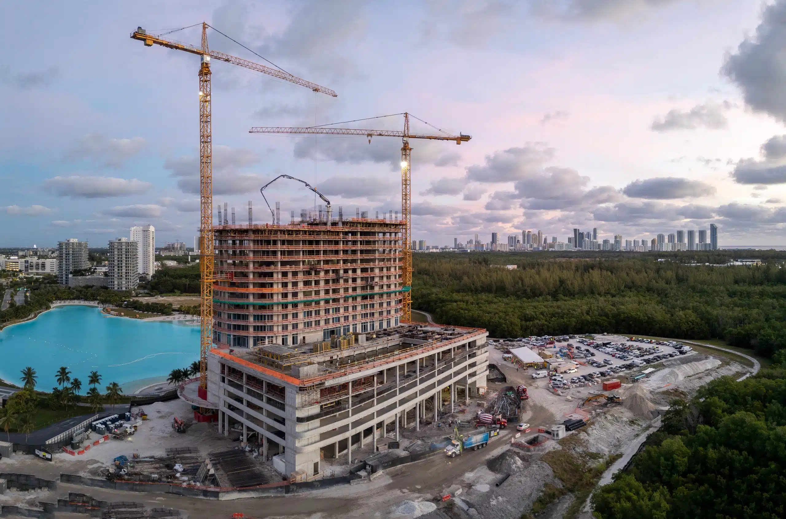 Aerial drone photo of One Park Tower under construction with two tower cranes at dusk, beside the turquoise SoLé Mia lagoon in North Miami, Florida