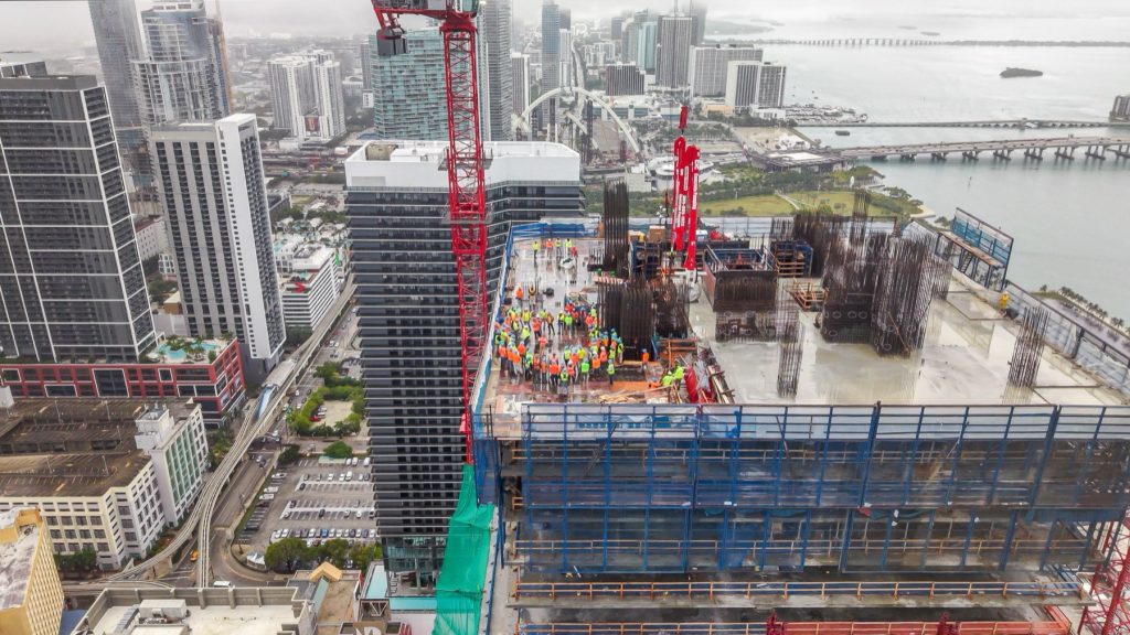 Drone view of a construction team assembled on the top deck of the Waldorf Astoria Miami tower during a topping out event, with downtown Miami and Biscayne Bay in the background