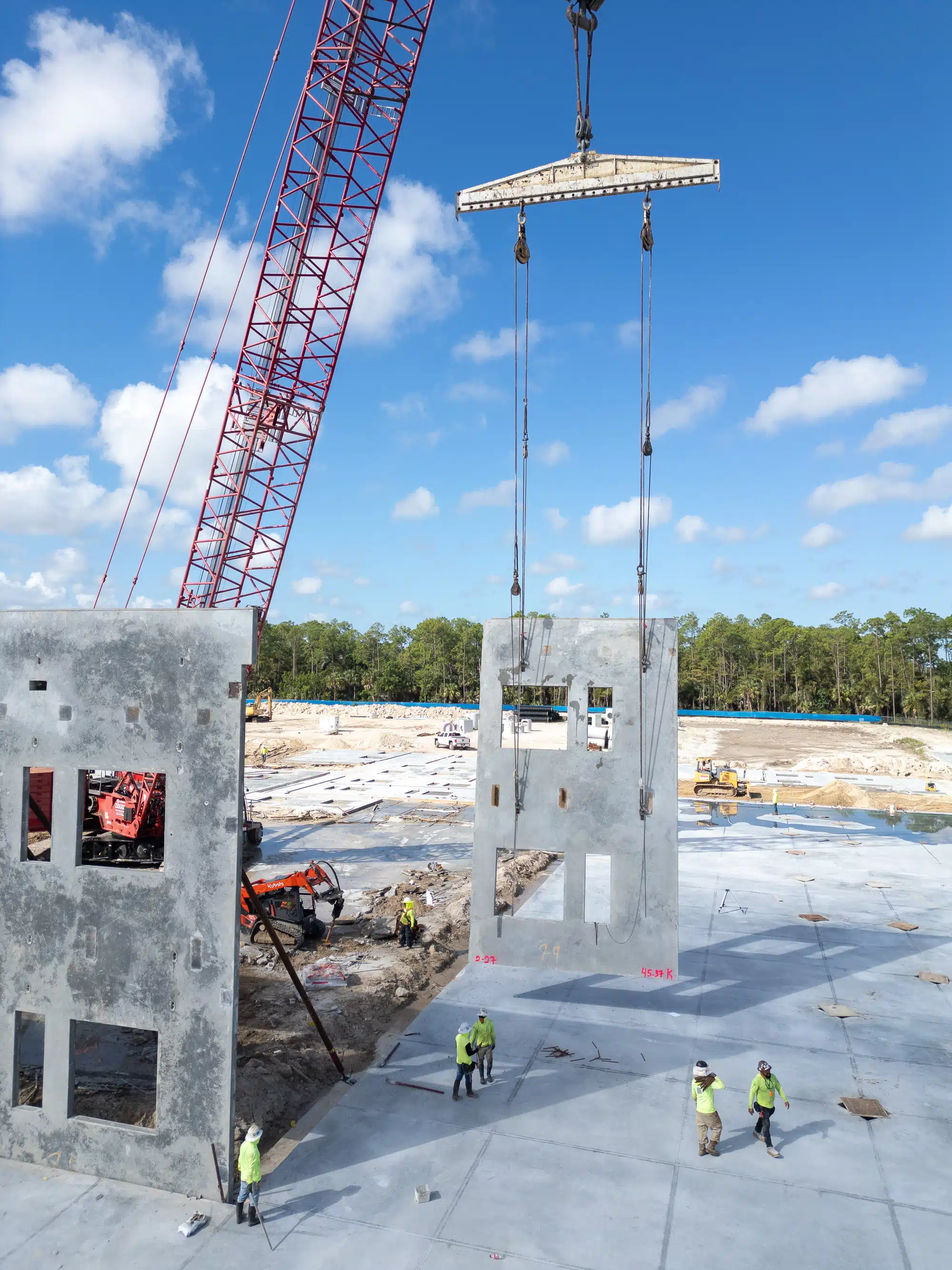 Drone photo of a crawler crane lifting a precast tilt-up concrete wall panel over a fresh slab while workers in safety vests guide the lift at the VB K-8 school site
