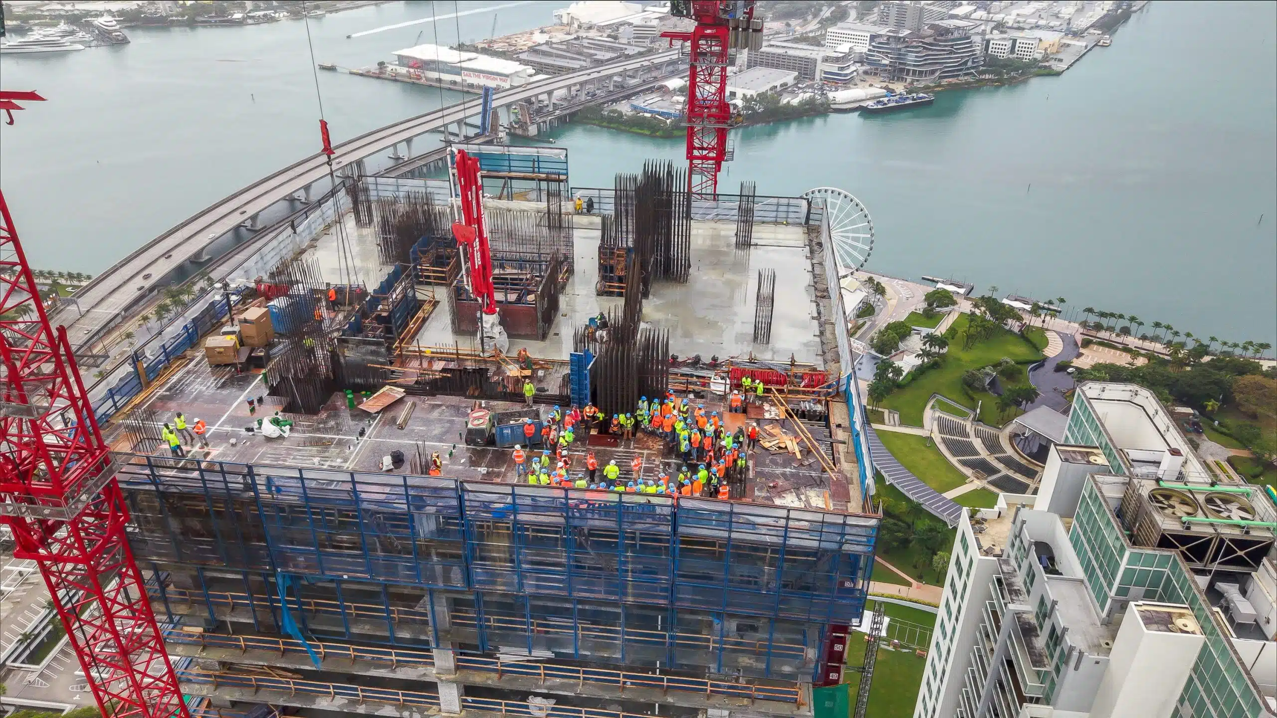 Close aerial view of construction crews gathered on the top deck of Waldorf Astoria Miami during a concrete pour milestone, with red tower cranes, Biscayne Bay and PortMiami behind