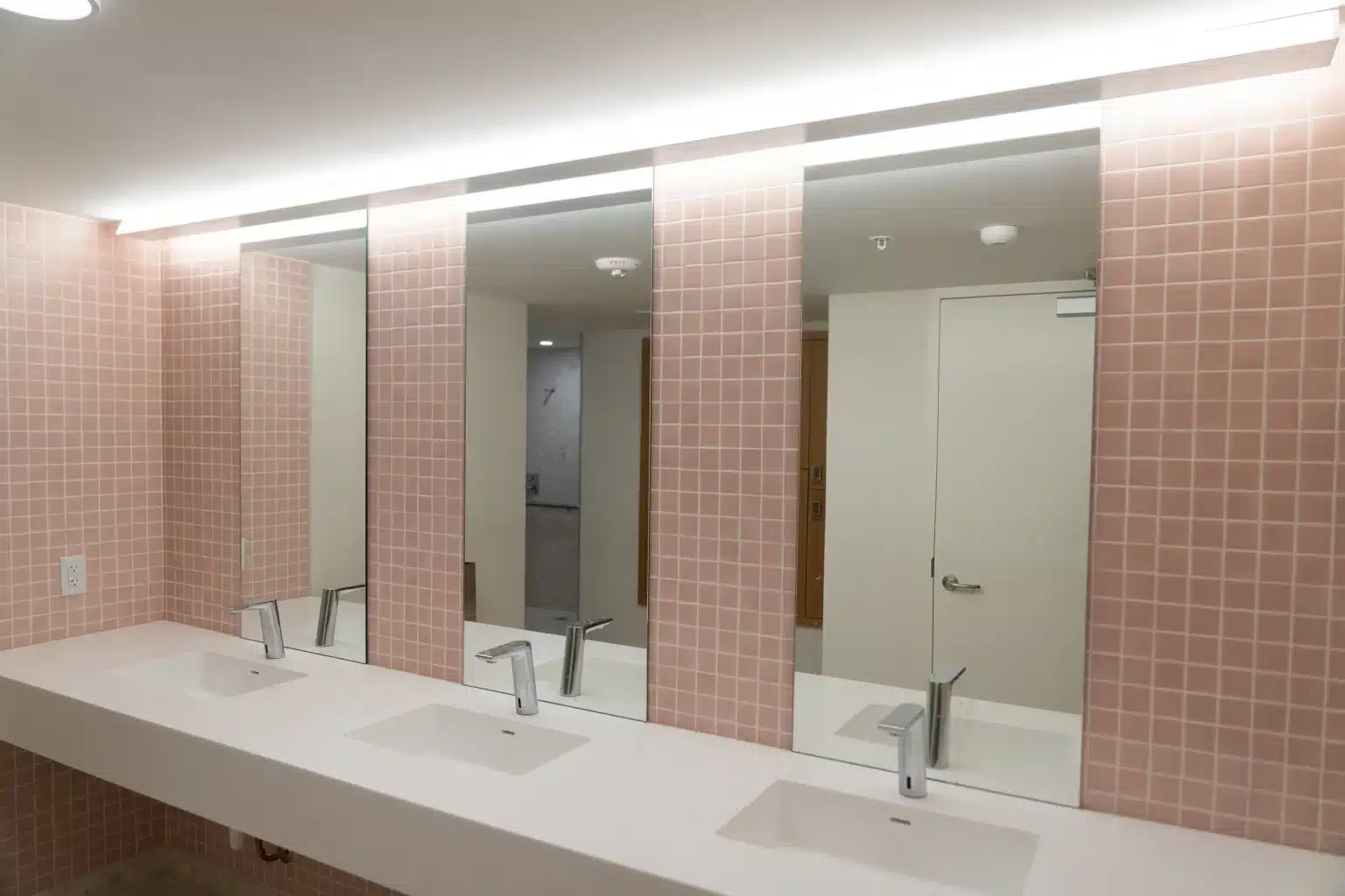 Amenity restroom vanity with pink square tile, solid surface counter and backlit mirrors at Flamingo Point in Miami Beach