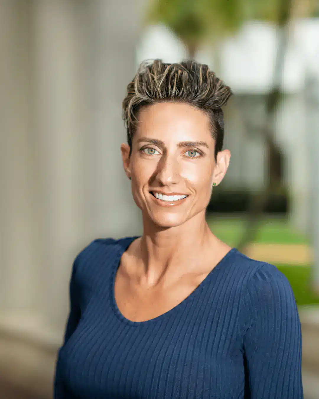 Outdoor professional headshot of a smiling woman with short styled hair in a navy top against a softly blurred background