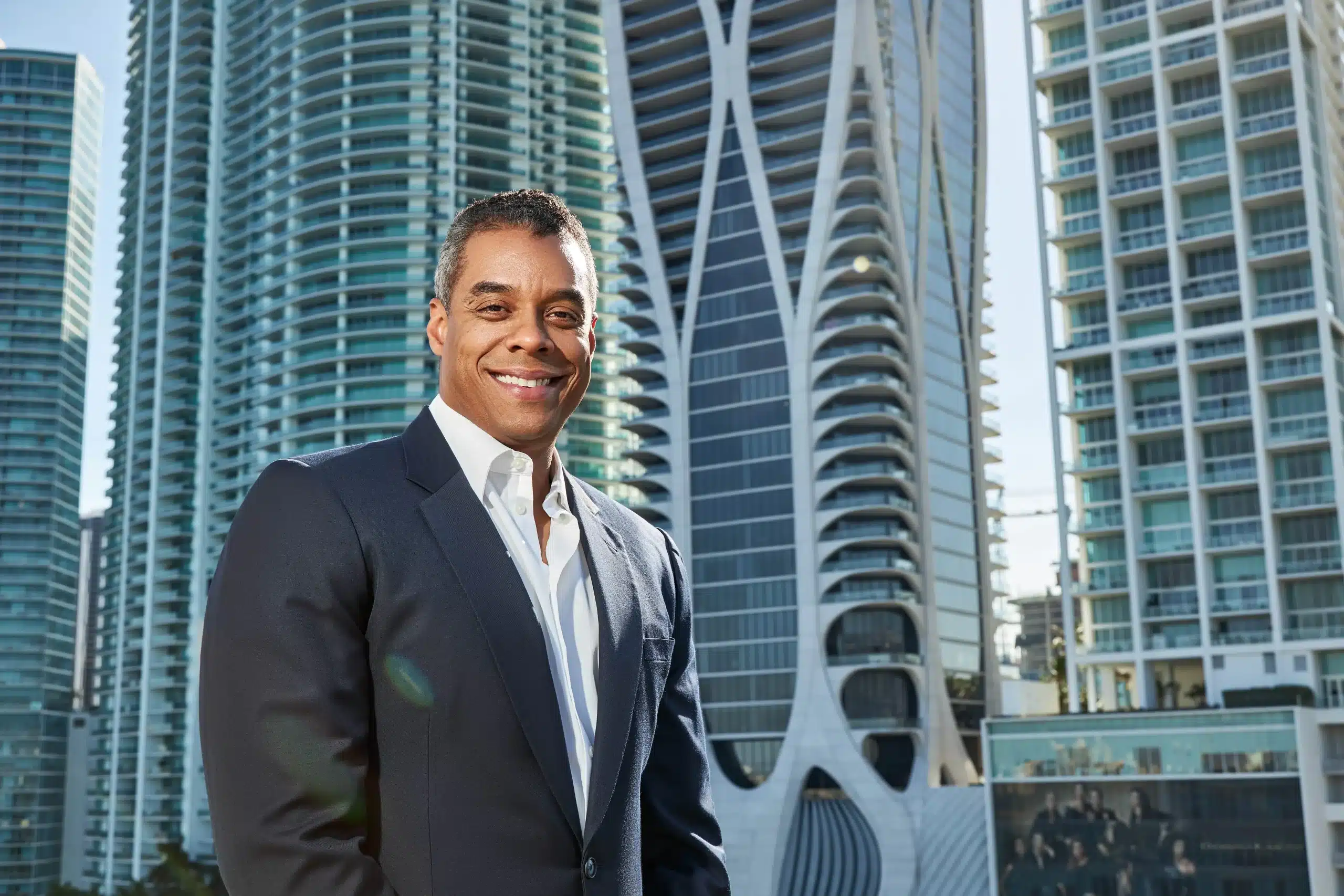 Executive portrait of a smiling man in a navy suit in front of the One Thousand Museum tower in downtown Miami